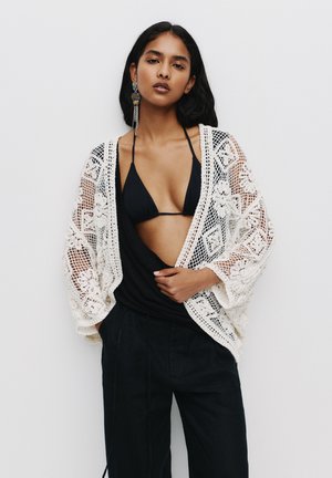 Creme-kleurige kanten cardigan met bloemenmotieven, gedragen over een zwart bikinitop en gedrapeerde zwarte stof, in combinatie met zwarte wijde broek.