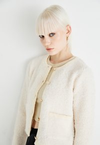 Krämfärgad cardigan med fluffig textur, rund halsringning och fem dekorativa knappar. Har två framfickor och en kort design.