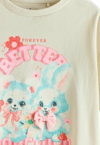 Lichtcrème t-shirt met een grafische print van twee cartoonkonijnen in blauw en roze, met een roze geruite strik en speelse tekst.