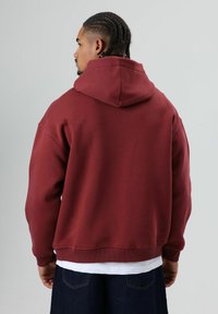 Donkerrode hoodie met een grote capuchon en geribbelde manchetten. Zachte stof, ontspannen pasvorm en een eenvoudig ontwerp. Draagt deze over een wit shirt en donkere spijkerbroek.