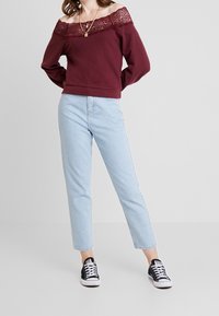 Sudadera burdeos sin mangas con escote de encaje, combinada con jeans rectos de color azul claro y zapatillas negras. Diseño simple; aspecto casual.