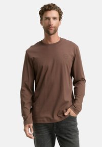Bruin overhemd met lange mouwen en een ronde hals, gemaakt van zacht materiaal, met een subtiel logo op de borst en een ontspannen pasvorm.