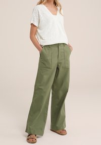 T-shirt léger en maille texturée blanche avec un col en V, associé à un pantalon large taille haute vert olive avec des poches avant. Sandales brunes.