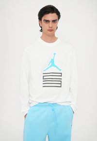 Camicia bianca a maniche lunghe con logo Jordan blu e testo nero. Indossata con pantaloni della tuta azzurri. Tessuto morbido, vestibilità casual.