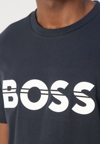 Marinblå bomulls t-shirt med en djärv vit "BOSS" logotyp och två horisontella ränder. Rund halsringning och mjuk textur.