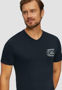 Marineblauwe v-hals t-shirt van katoen, met een kleine witte grafische print op de linkerkant van de borst met tekst en visgerelateerde afbeeldingen.