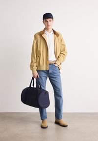 Beige bomberjakke med brun krave, hvid polo skjorte, blå jeans og lysebrune støvler. Holder en mørkeblå duffeltaske med to håndtag.