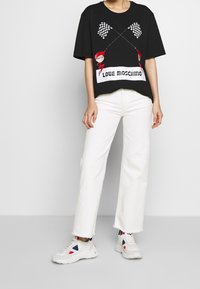 T-shirt oversize nera con stampa grafica di bandiere a scacchi e personaggio dei cartoni animati, abbinata a jeans bianchi a gamba larga e sneakers bianche.