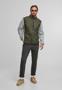 Chaleco acolchado en verde oliva con mangas grises, cierre de cremallera en el frente, bolsillos laterales y detalles de costura blanca, combinado con pantalones oscuros y zapatos beiges.