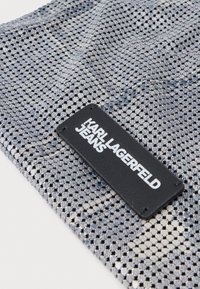 Zilverkleurige gaasstof met een zwart rechthoekig label erop gestikt, met de tekst "KARL LAGERFELD JEANS" in witte, vetgedrukte letters.