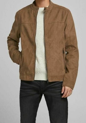 Homme portant une veste en daim brun clair sur un pull en maille blanc et un pantalon noir, debout avec une main dans la poche et l'autre reposant sur le côté.