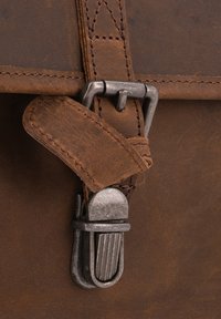 Nahbrown lædertaske i close-up med en metalspænde, syet rem og struktureret overflade. Detaljer inkluderer en buet, sølvfarvet spænde og glat læder.
