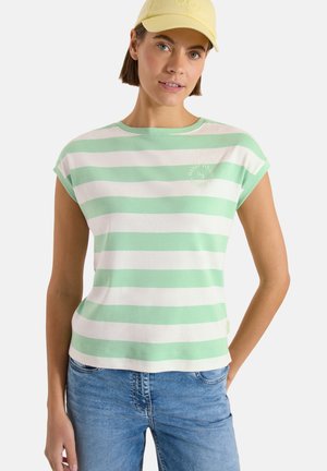 Jeune femme portant un débardeur rayé vert et blanc, un jean bleu et une casquette jaune clair, posant avec une main dans la poche.