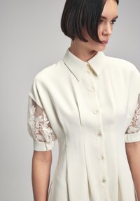 Witte jurk met een knopenlijst aan de voorkant, klassieke kraag en korte pofmouwen, versierd met ingewikkelde kantborduursels. Zacht, glad materiaal.