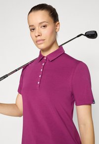 Lila Polohemd mit Kragen, mit Knopfverschluss und kurzen Ärmeln. Ein schwarzer Golfschläger ruht auf der Schulter und fügt Kontext hinzu.