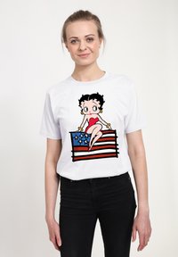 Hvid t-shirt med et grafisk motiv af en tegneseriefigur, der sidder på et stiliseret amerikansk flag i røde, hvide og blå farver.