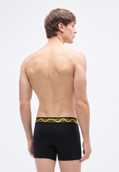 Μαύρα μπόξερ εσώρουχα με διακοσμητική ζώνη μέσης σε χρυσό σχέδιο, απεικονιζόμενα από πίσω. Το υλικό φαίνεται λείο και εφαρμοστό.