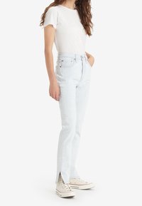 Jeans de mezclilla azul claro con cintura alta, corte recto, aberturas laterales en el dobladillo, complementados con una camiseta blanca de manga corta y zapatillas beige.