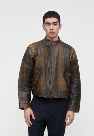 Jeune homme aux cheveux noirs courts portant une veste en cuir marron usée avec une fermeture éclair asymétrique et un pantalon noir, debout devant un fond uni.