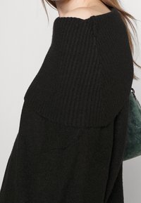 Svart stickad tröja med ribbad krage och axeldesign. Mjuk textur, med diagonala sömmar och en avslappnad passform.