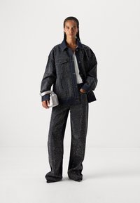 Chaqueta de denim y pantalones en azul oscuro con un patrón salpicado metálico. La chaqueta tiene bolsillos frontales y un gran cuello. Lleva un bolso pequeño plateado.
