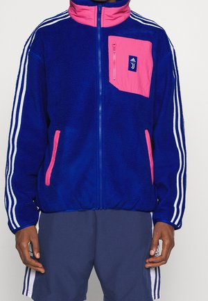 Blaue Fleecejacke mit rosa Akzenten, Reißverschlusstasche auf der Brust, drei weiße Streifen an den Ärmeln, getragen mit marineblauen Shorts mit weißen Streifen.
