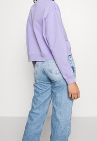 Person i ljuslila kort tröja och högmidjade ljusblå jeans, stående mot en enfärgad ljus bakgrund.