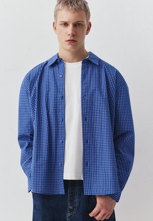 Mladý muž s krátkými blond vlasy, který nosí modrou kostkovanou košili přes bílé tričko a tmavé džíny, stojící u prosté bílé stěny.