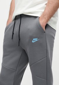 Άνδρας που φοράει γκρι αθλητική φόρμα Nike με μαύρα κορδόνια και τα χέρια στις τσέπες, ορατός από τη μέση έως το μέσο μηρού.