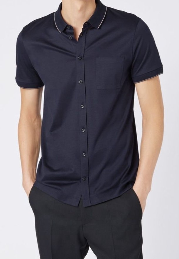 Homme portant une chemise bleu marine à manches courtes avec une poche poitrine et un pantalon noir, mains dans les poches.