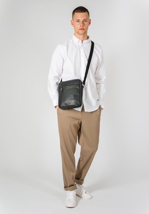 Borsa a tracolla in pelle nera con tasca con zip, completata da una tracolla testurizzata, appoggiata su un modello che indossa una camicia bianca e pantaloni beige.