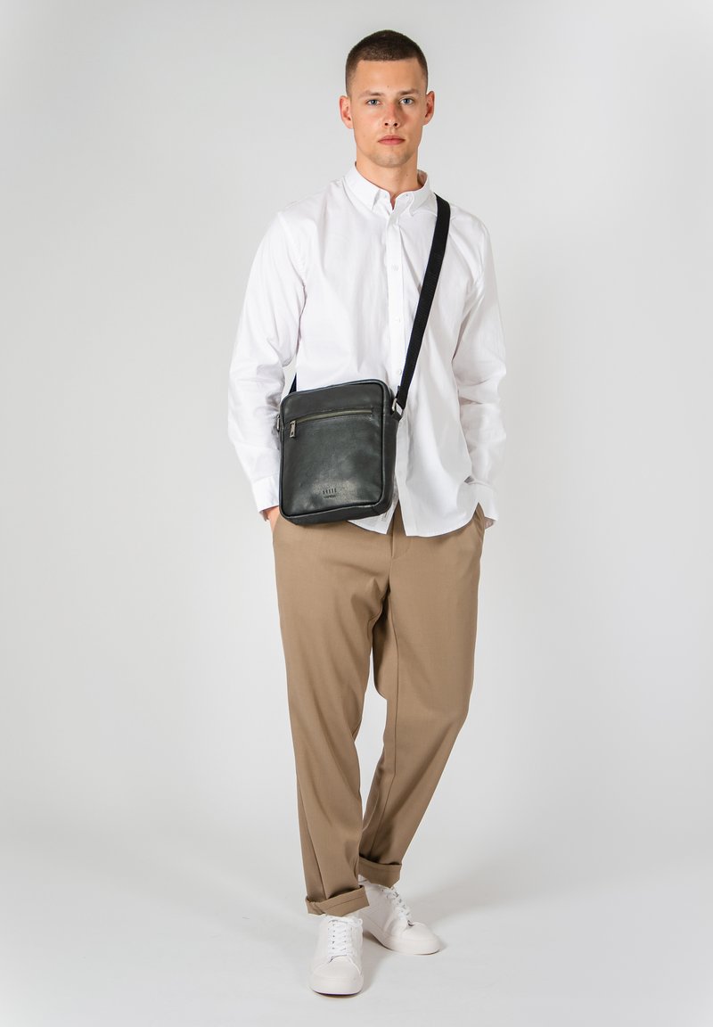 Černá kožená crossbody taška se zipovou kapsou, doplněná strukturovaným popruhem, odpočívající na modelce v bílé košili a béžových kalhotách.