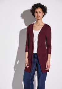 Bordeaux lange cardigan met een diepe V-hals, lange mouwen, open voorkant, gecombineerd met een witte top en blauwe jeans. Gladde textuur, casual ontwerp.