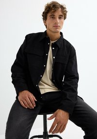 Camisa negra de botones con dos bolsillos en el pecho, combinada con una camiseta de cuello redondo beige claro y jeans oscuros, sentado en un taburete.