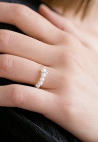 Zlatý prsteň s malými bielymi perlami a jedným akcentom podobným diamantu, umiestnený na ruke položené na tmavom textilnom pozadí.