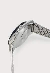 Montre-bracelet en métal argenté avec une lunette noire et un bracelet en maillons texturés en métal argenté, présentée de profil et au dos.