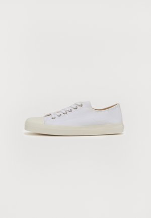 Zapatilla de lona blanca con punta redonda, suela plana de goma crema, seis ojetes para cordones y sutiles detalles de costura en la parte superior.