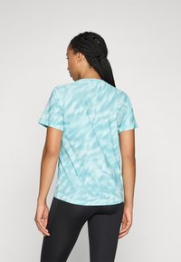 Donna con capelli intrecciati che indossa una maglietta a maniche corte azzurro chiaro a fantasia e leggings neri, vista di spalle su uno sfondo neutro.