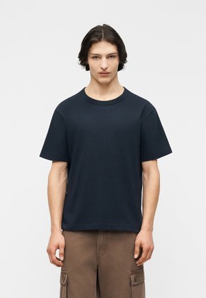 Mladý muž so stredne dlhými tmavými vlasmi, oblečený v jednoduchom tmavomodrom tričku a hnedých vojenských nohaviciach, na jednoduchom pozadí.