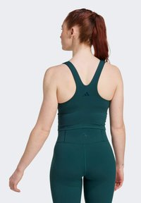 Zeleni sportski grudnjak s dizajnom racerback, izrađen od rastezljivog materijala. Ima suptilan logotip brenda na leđima, uparen s odgovarajućim tajicama.