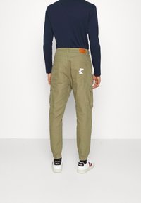 Oliwkowe spodnie cargo z elastycznymi mankietami, posiadające dwie boczne kieszenie oraz tylnią naszywkę z białym logo na brązowej skórzanej metce.