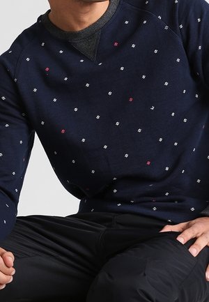 Mann trägt marineblauen Pullover mit kleinen weißen und roten Sternmustern über dunklem grauem Unterhemd, sitzt mit schwarzen Hosen.