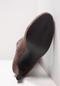 Botín de ante marrón hasta el tobillo con un tacón afinado y una suela de goma negra texturizada. Detalle de cremallera lateral, superficie superior lisa y sin patrones visibles.