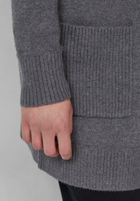 Pull en maille gris avec des poignets côtelés et un corps texturé, doté d'une poche latérale. Le matériau semble doux et chaud, idéal pour le temps froid.