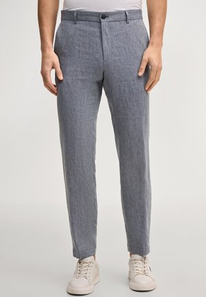 Uomo con pantaloni aderenti grigio chiaro e scarpe da ginnastica bianche, in piedi contro uno sfondo chiaro e uniforme.