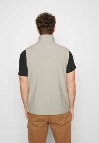 Ljust beige fleeceväst med hög krage, bakvy. I kombination med en svart kortärmad skjorta, minimalistisk design, mjuk textur, inga synliga knäppningar.