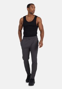 Atletische man in zwarte tanktop, donkergrijze broek en zwarte sneakers die loopt en naar links kijkt tegen een witte achtergrond.