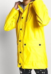 Żółta wodoodporna kurtka z czarnymi guzikami i kapturem ze sznurkiem. Posiada przednią kieszeń oraz wewnętrzną podszewkę w paski.