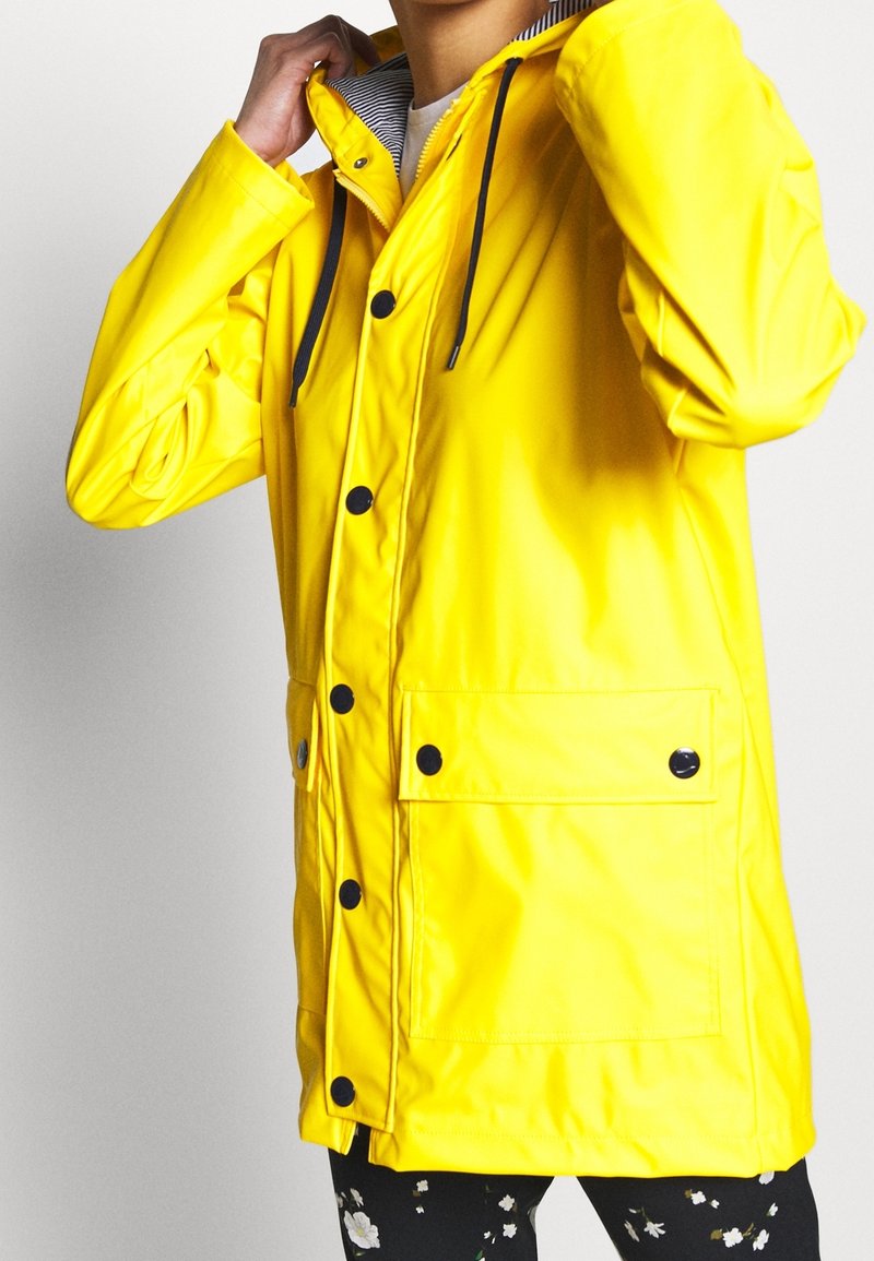 Żółta wodoodporna kurtka z czarnymi guzikami i kapturem ze sznurkiem. Posiada przednią kieszeń oraz wewnętrzną podszewkę w paski.