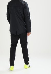 Zwarte sportjas van lichtgewicht stof met een gladde textuur. Zwarte broek met witte zijstrepen en felgele sportschoenen.